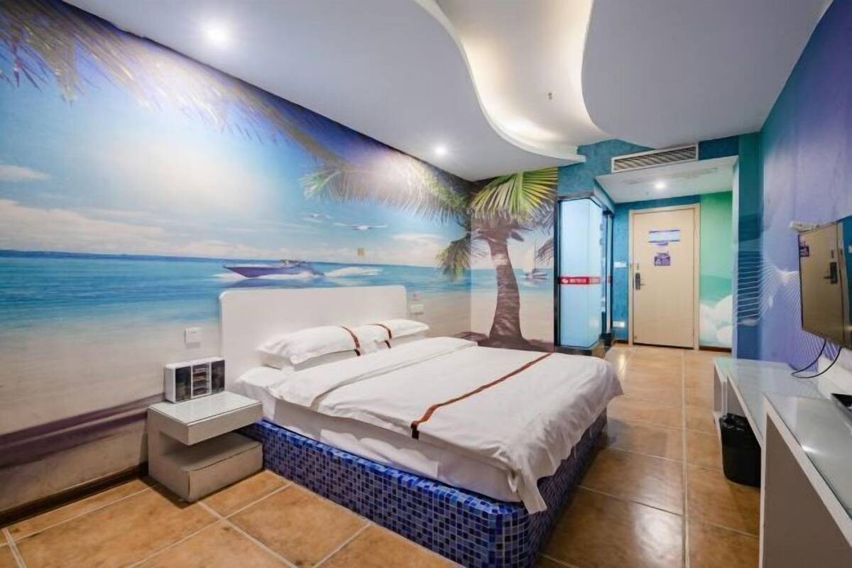 A hotel room with a beach mural, a bed with white linens, a nightstand, and a bathroom door.