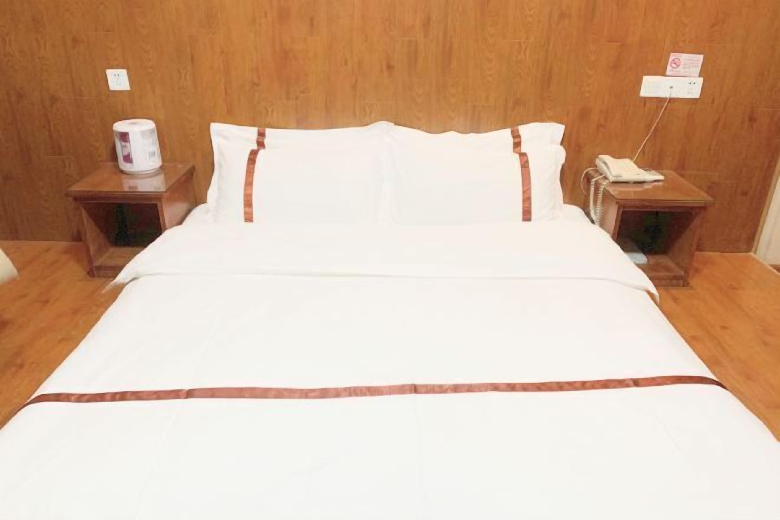 A neatly made bed with white bedding and brown accents, two bedside tables with a telephone and a lamp, and a wooden wall.