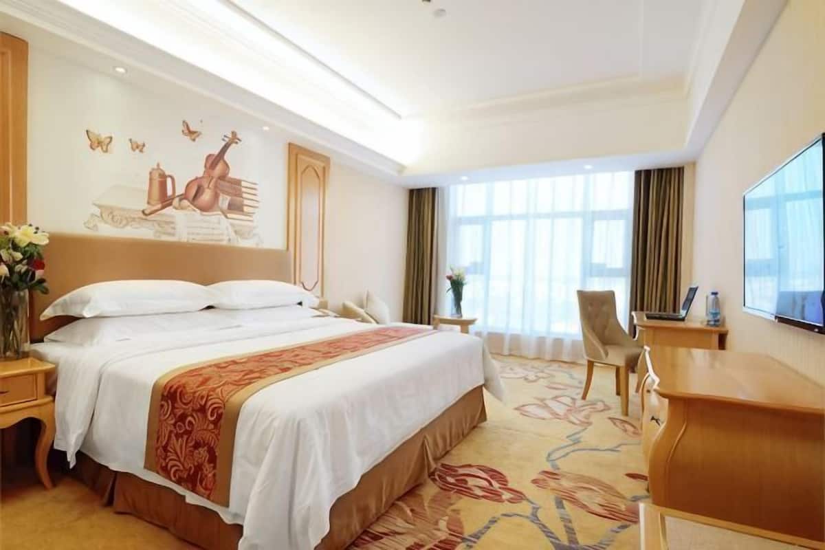 A hotel room with a large bed, a desk, a chair, a TV, and a window with curtains.