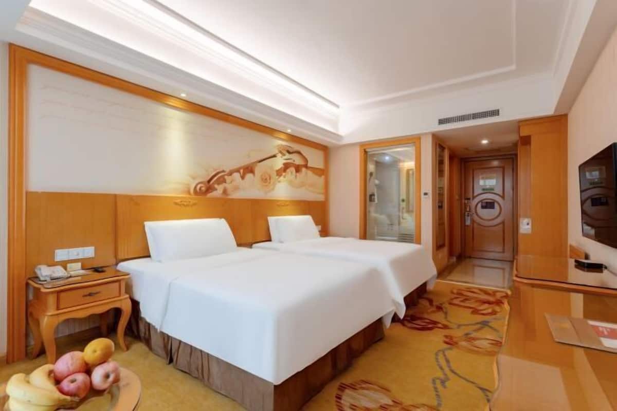 A hotel room with two beds, a wooden headboard, a painting, a small table with a phone, and a fruit bowl.
