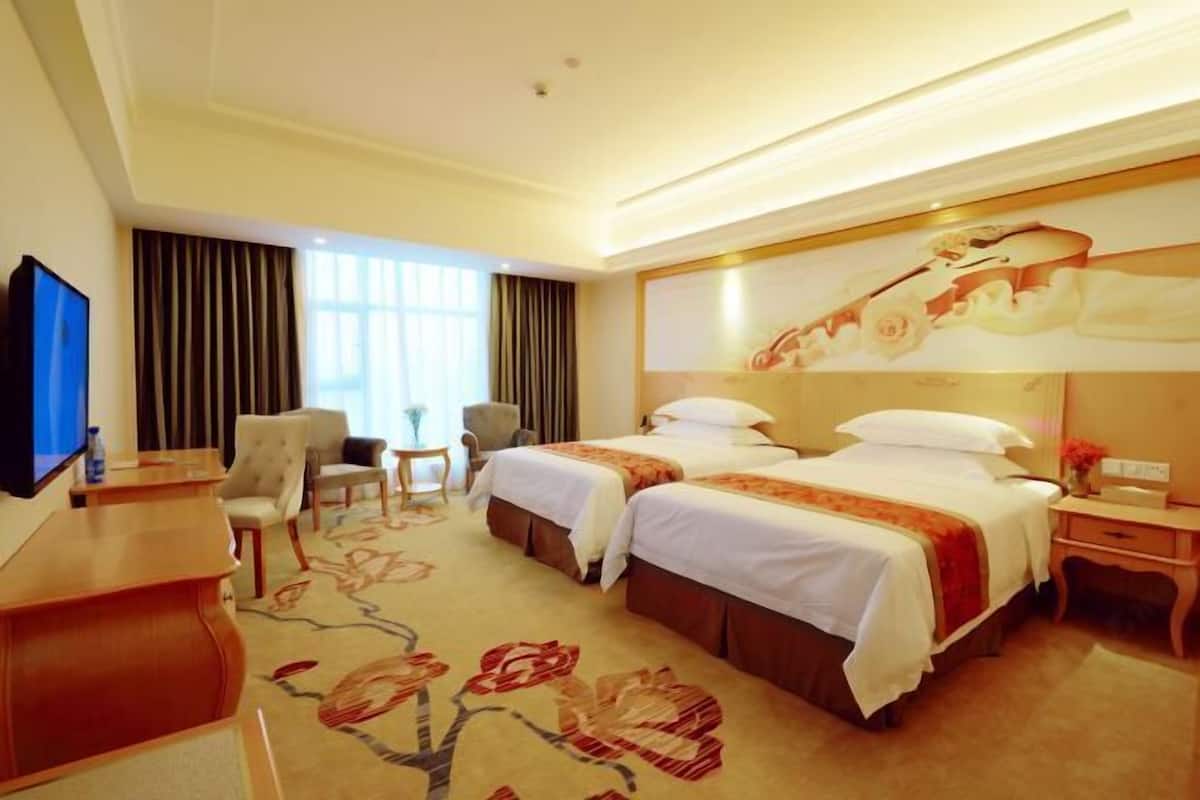 A hotel room with two beds, a desk, a chair, a TV, and a large window with curtains.