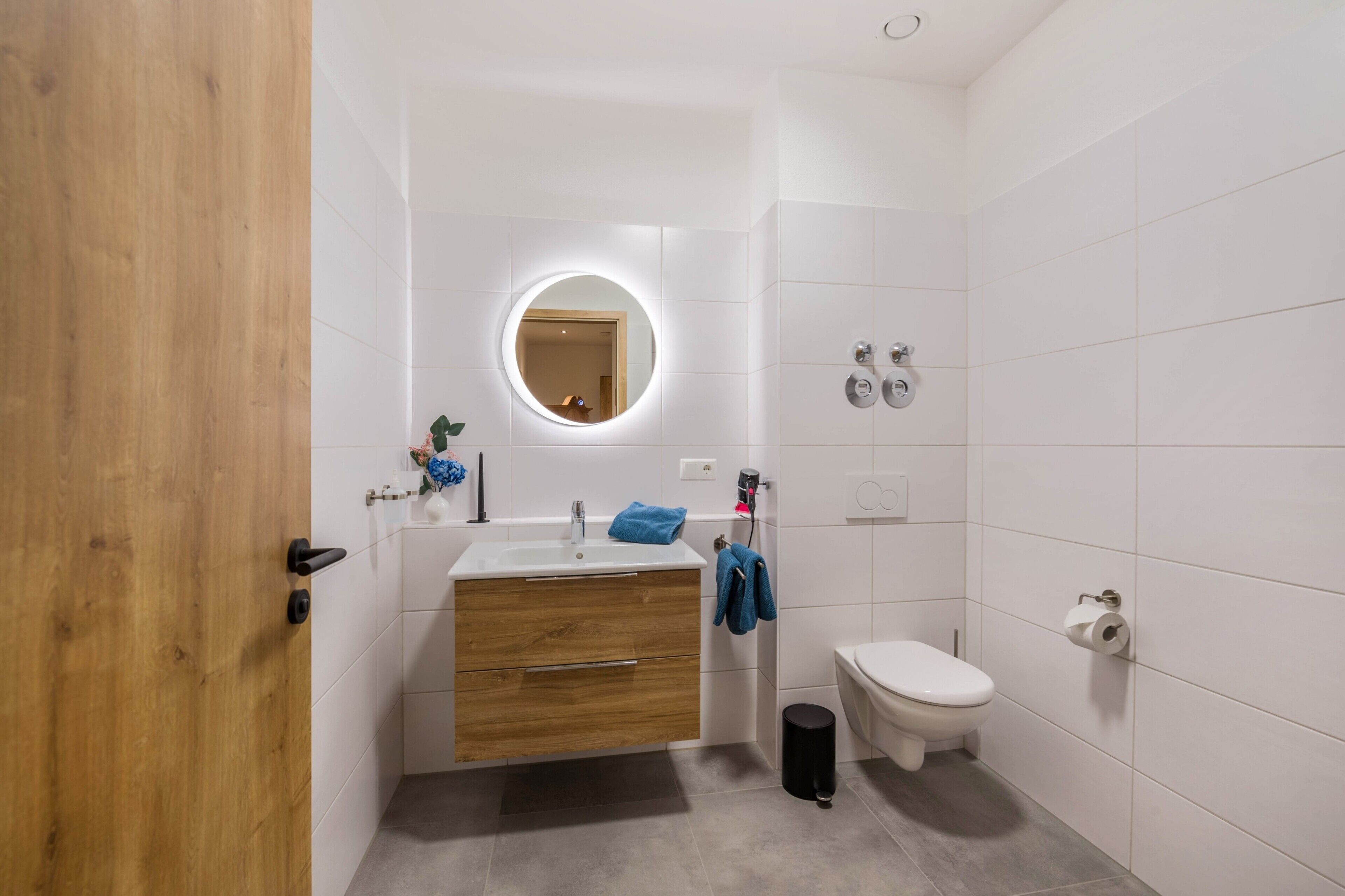 Ein Badezimmer mit Holztür, rundem Spiegel, Waschbecken, Toilette und Mülleimer.