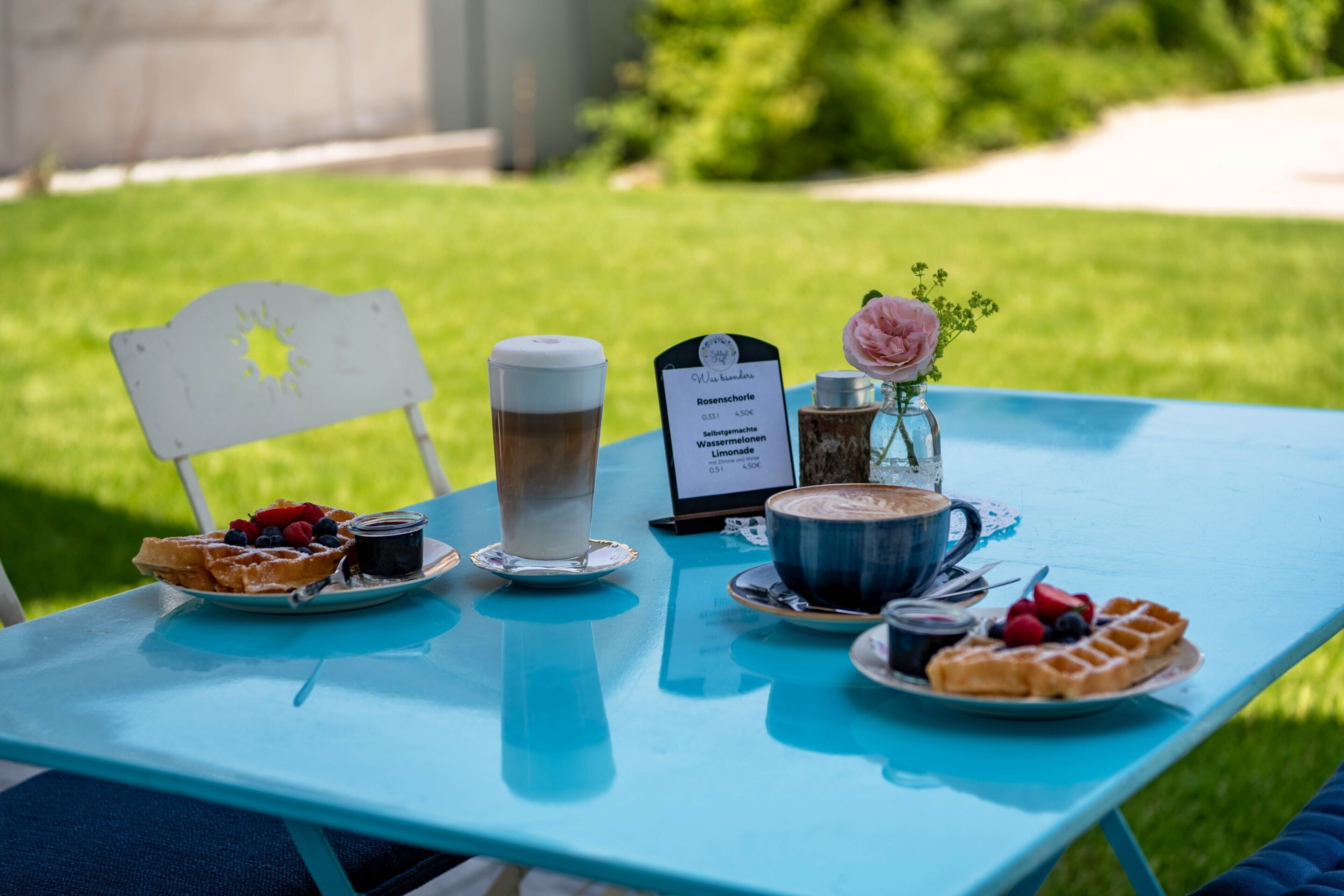 Ein Tisch mit Frühstücksangeboten, darunter Waffeln, Kaffee und ein Getränk, draußen mit einer Speisekarte.