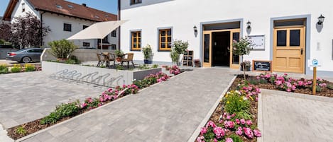Un bâtiment à la façade blanche, doté de nombreuses fenêtres et d’un espace d’entrée pavé agrémenté de parterres de fleurs.