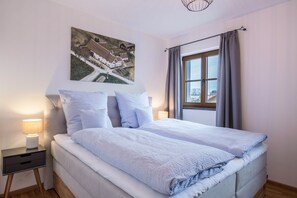 A bedroom with a bed, a nightstand, a lamp, a window with curtains, and a painting on the wall.