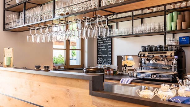 Een café interieur met een houten toonbank, opgeschorte wijnglazen, een koffieapparaat en een krijtbord met de menukaart.