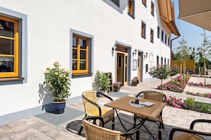 A building with a white exterior, yellow-framed windows, outdoor seating with wicker chairs, and potted plants.