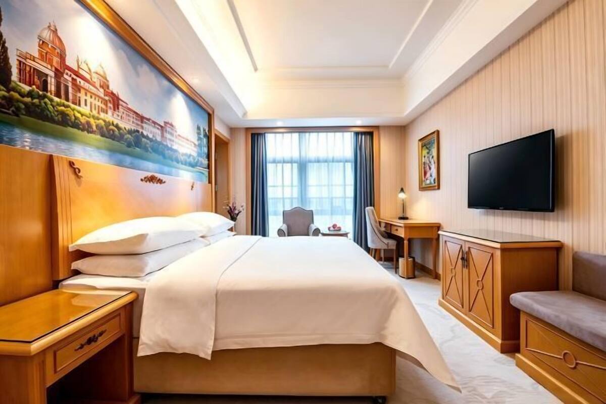 A hotel room with a bed, a desk, a TV, and a painting on the wall.