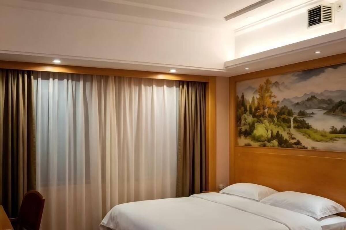 A hotel room with a bed, a painting on the wall, and a window with curtains.