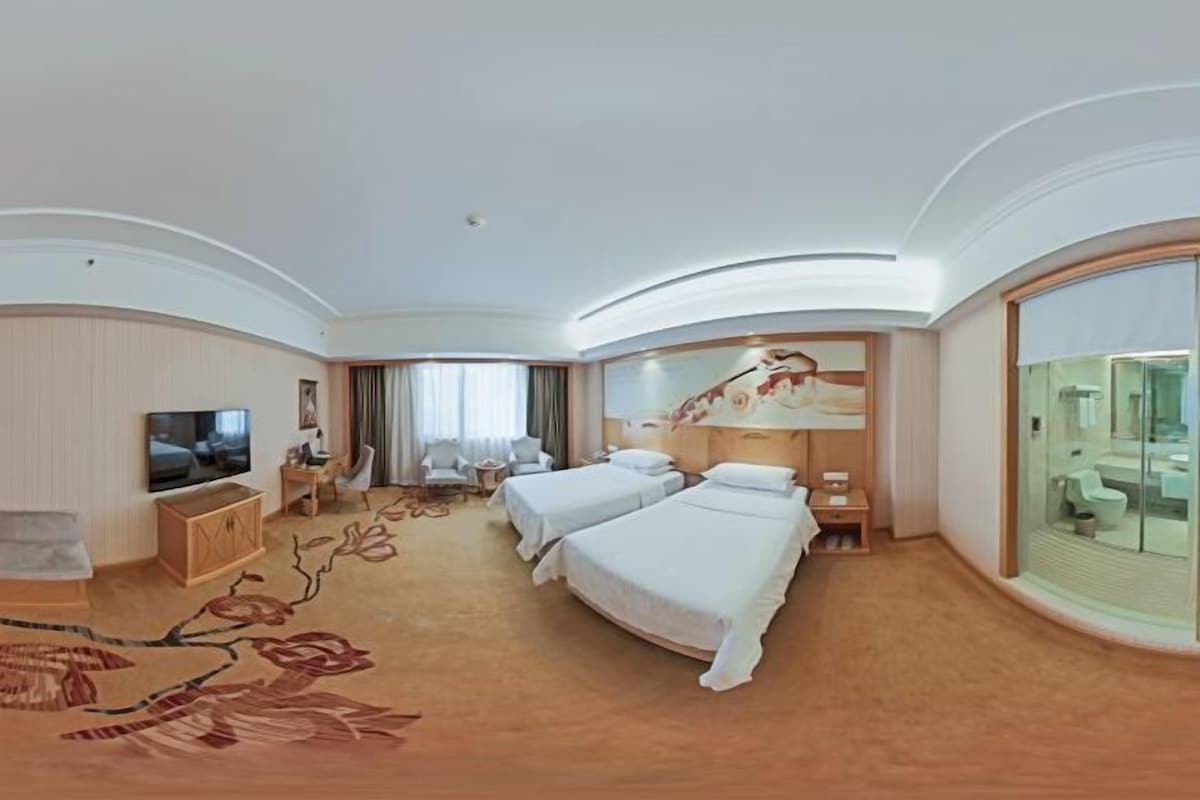 Ein Hotelzimmer mit zwei Betten, einem Fernseher, einem Schreibtisch, einem Sessel und einem großen Fenster.