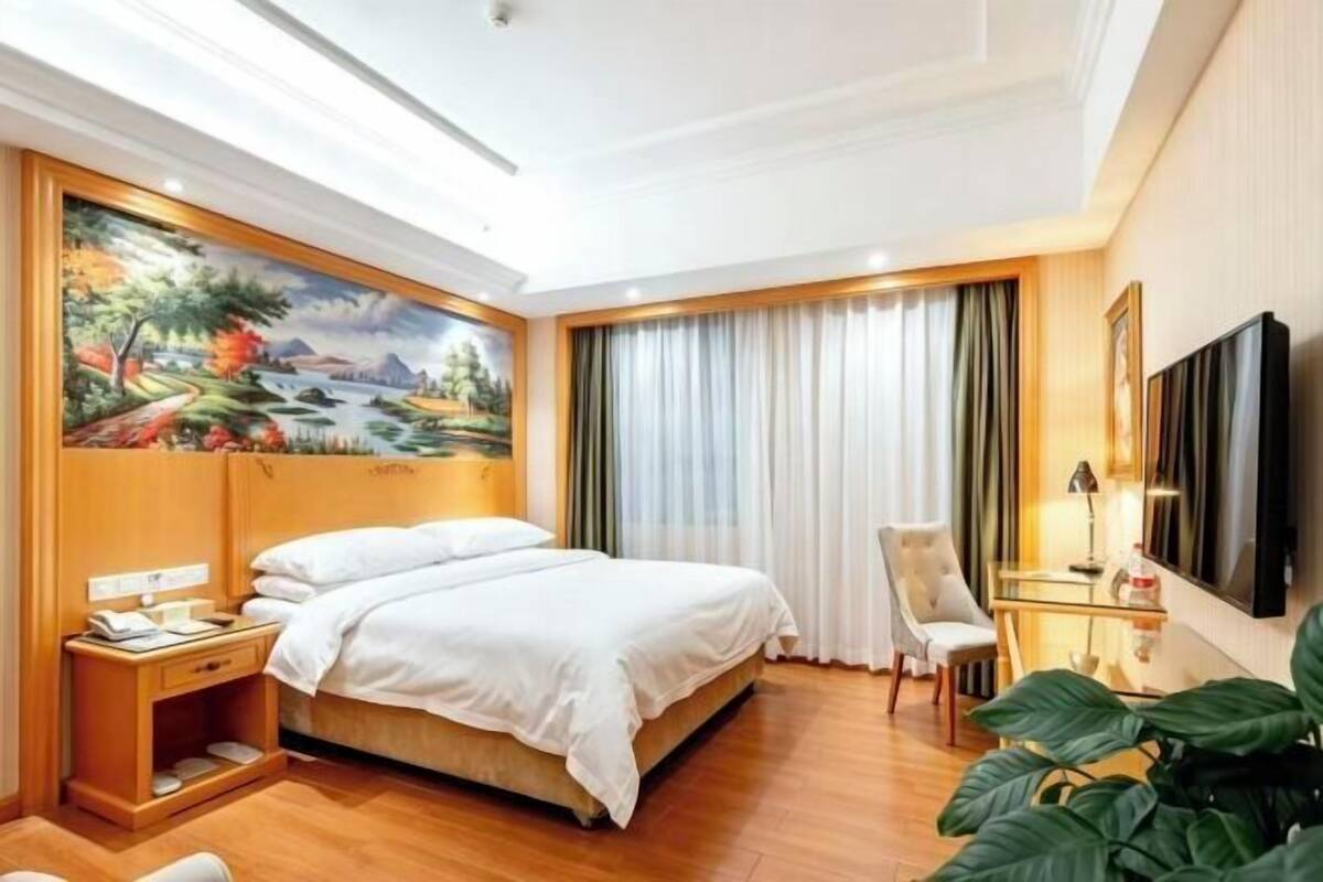 A hotel room with a bed, a desk, a chair, a TV, and a painting on the wall.
