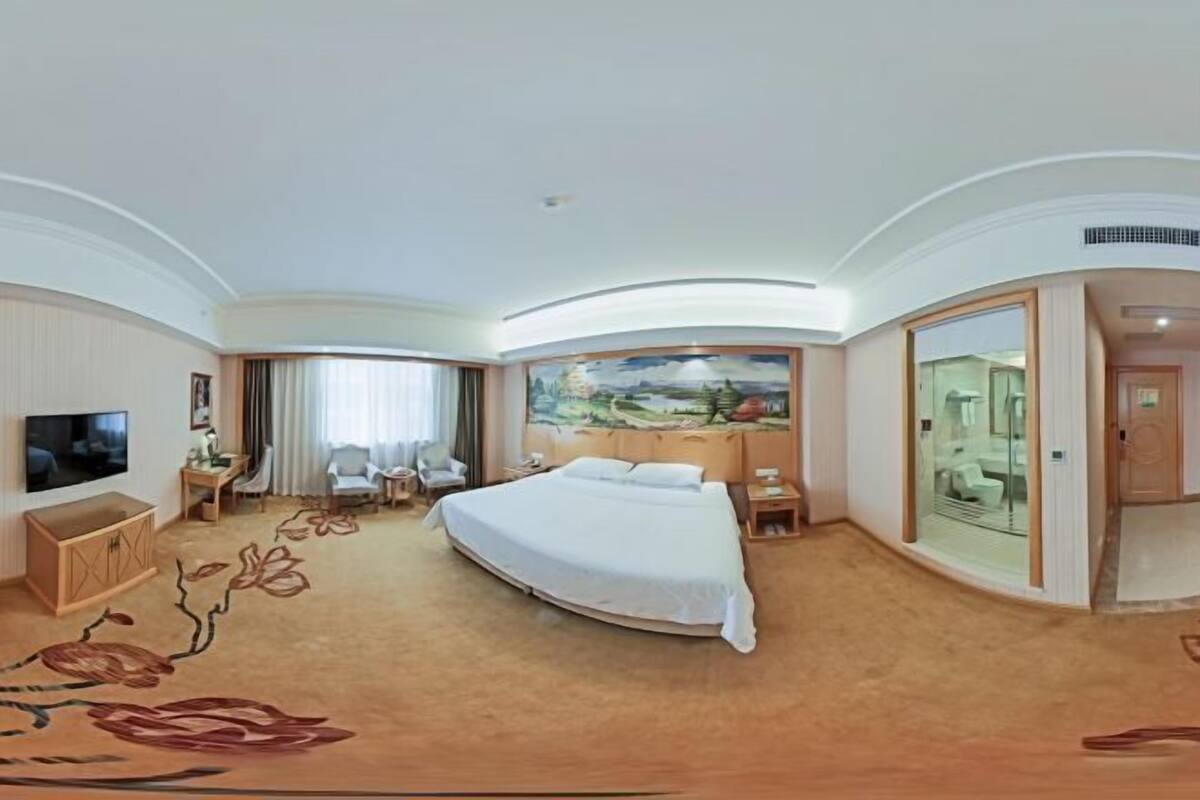 A hotel room with a bed, a TV, a desk, a chair, and a painting on the wall.