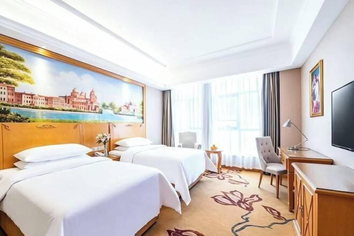 A hotel room with two beds, a desk, a chair, a TV, and a painting on the wall.