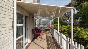 A covered outdoor area with a table and chairs, a white fence, and lush greenery in the background.
