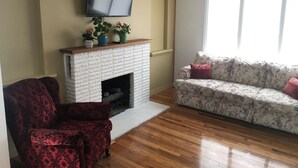 A living room with a fireplace, a television mounted above it, a red patterned armchair, a floral sofa, and a large window providing natural light. - Classic pretty home with nice patio in Rigby. (Rigby)