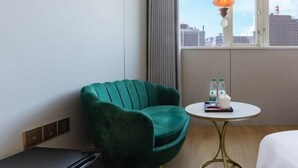Habitación de hotel con una silla verde, una mesita redonda con tetera y agua embotellada, y vistas de la ciudad a través de un ventanal grande.