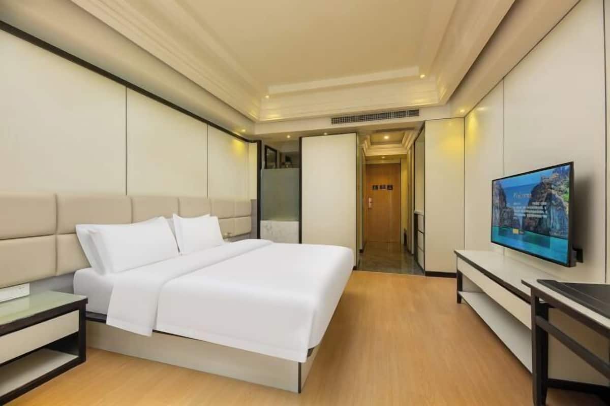 A modern hotel room with a bed, bedside table, TV, and a hallway leading to another room.