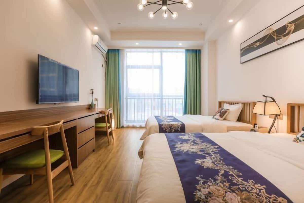 A hotel room with a wooden desk, two beds with floral bedspreads, a television, and a large window with curtains.
