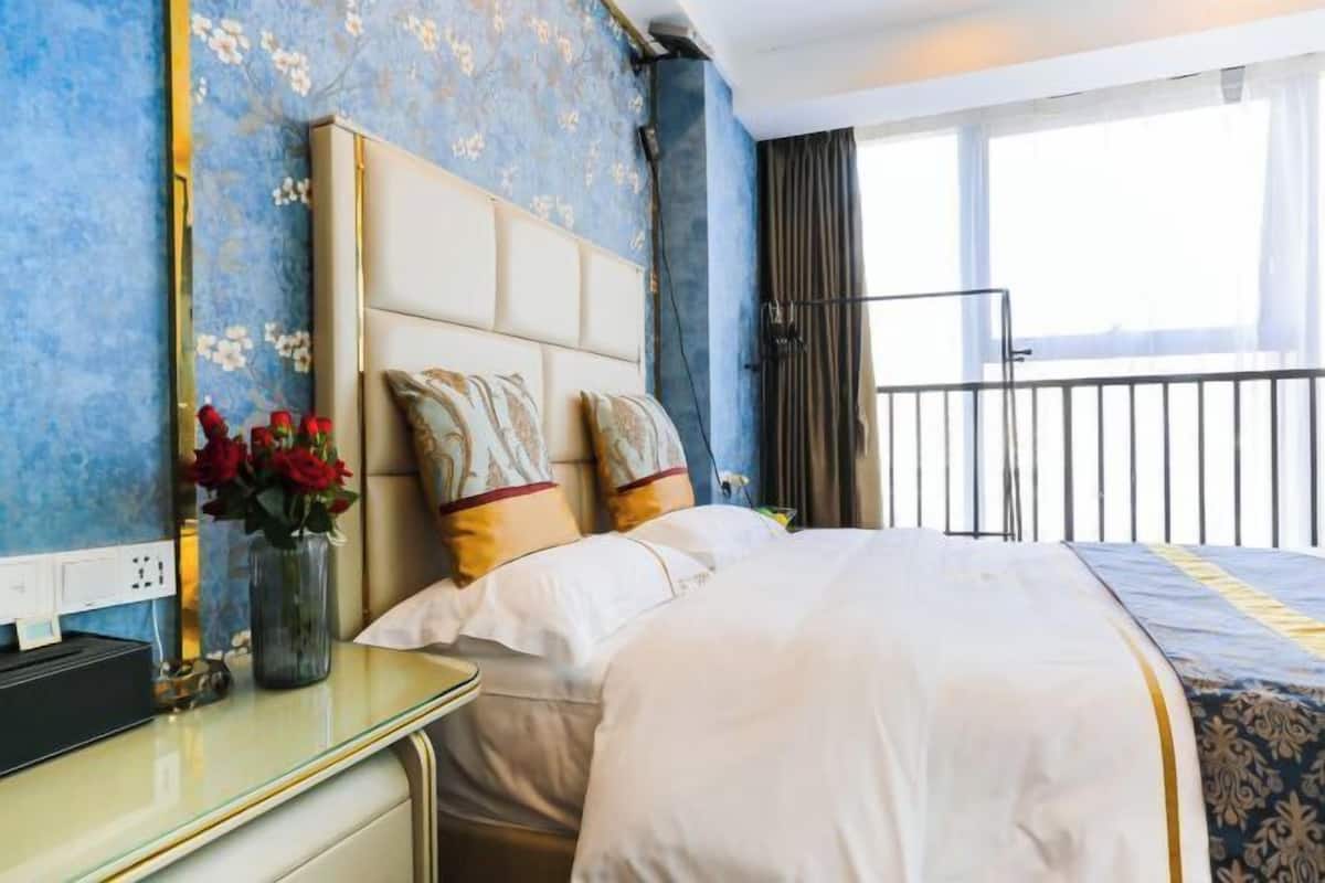 A hotel room with a bed, a bedside table with a vase of flowers, a window with a view, and a balcony railing.