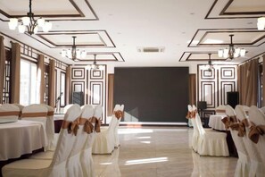 Una sala da banchetti con file di tavoli e sedie, uno schermo grande e lampadari.