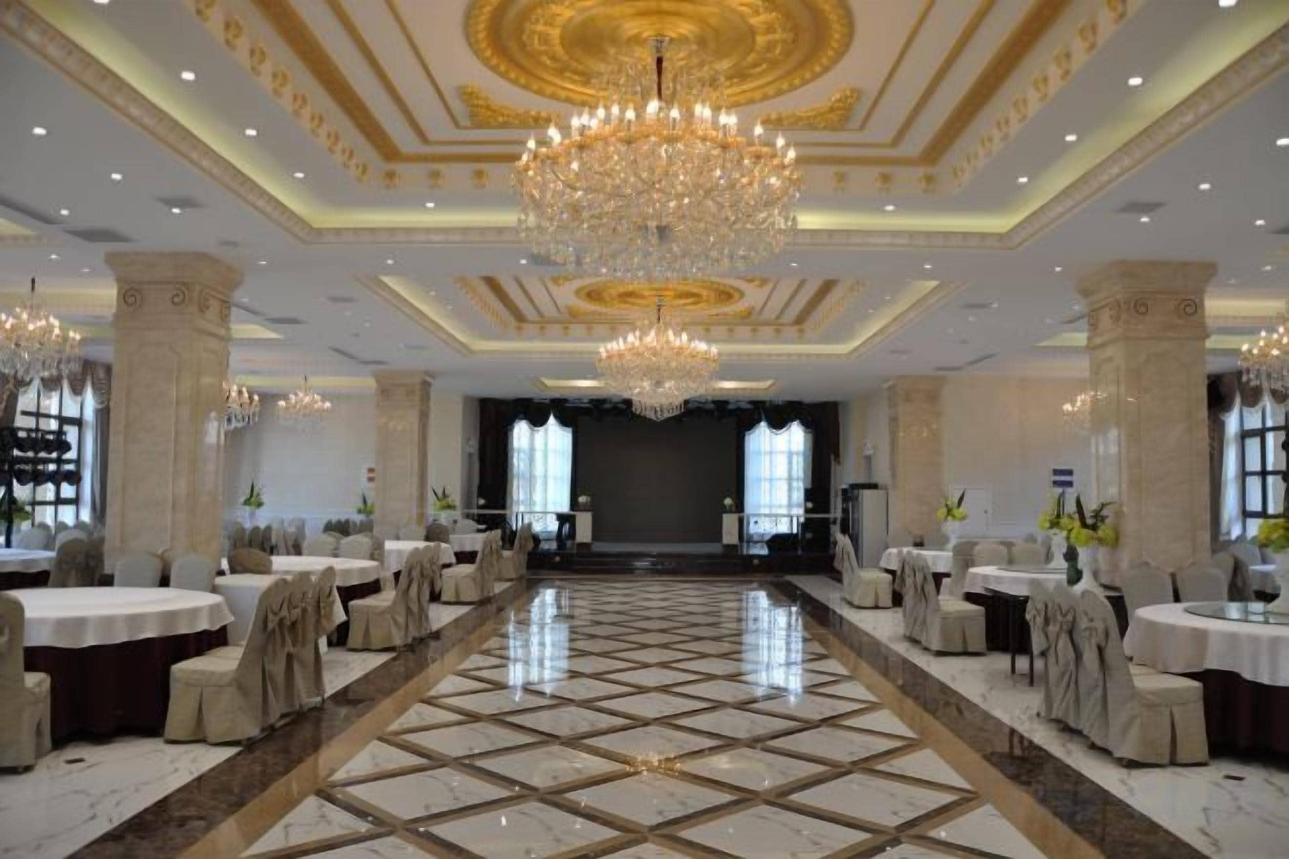 Un amplio salón de banquetes con un escenario central, candelabros y suelo de mármol.