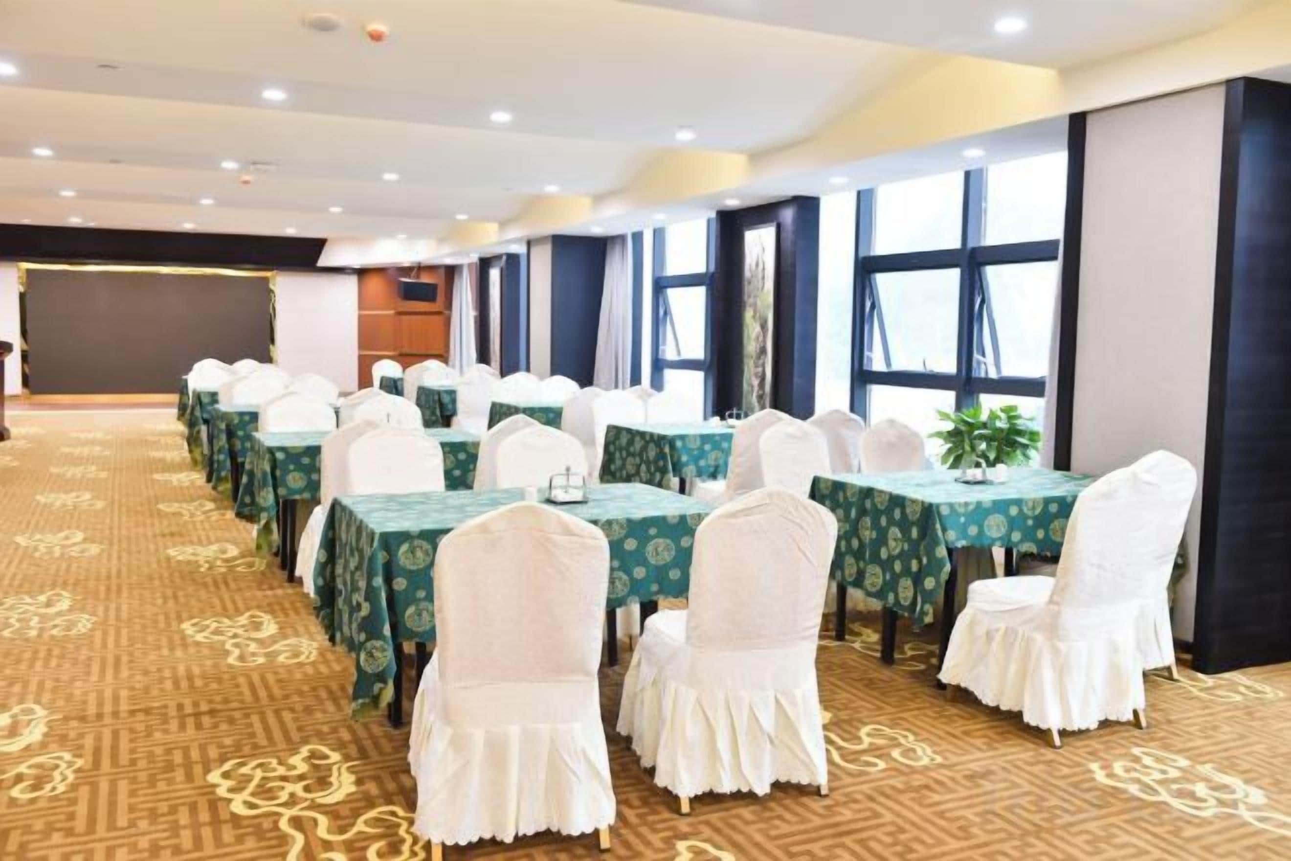 Un salón de conferencias con filas de mesas y sillas, suelo alfombrado y ventanas amplias.