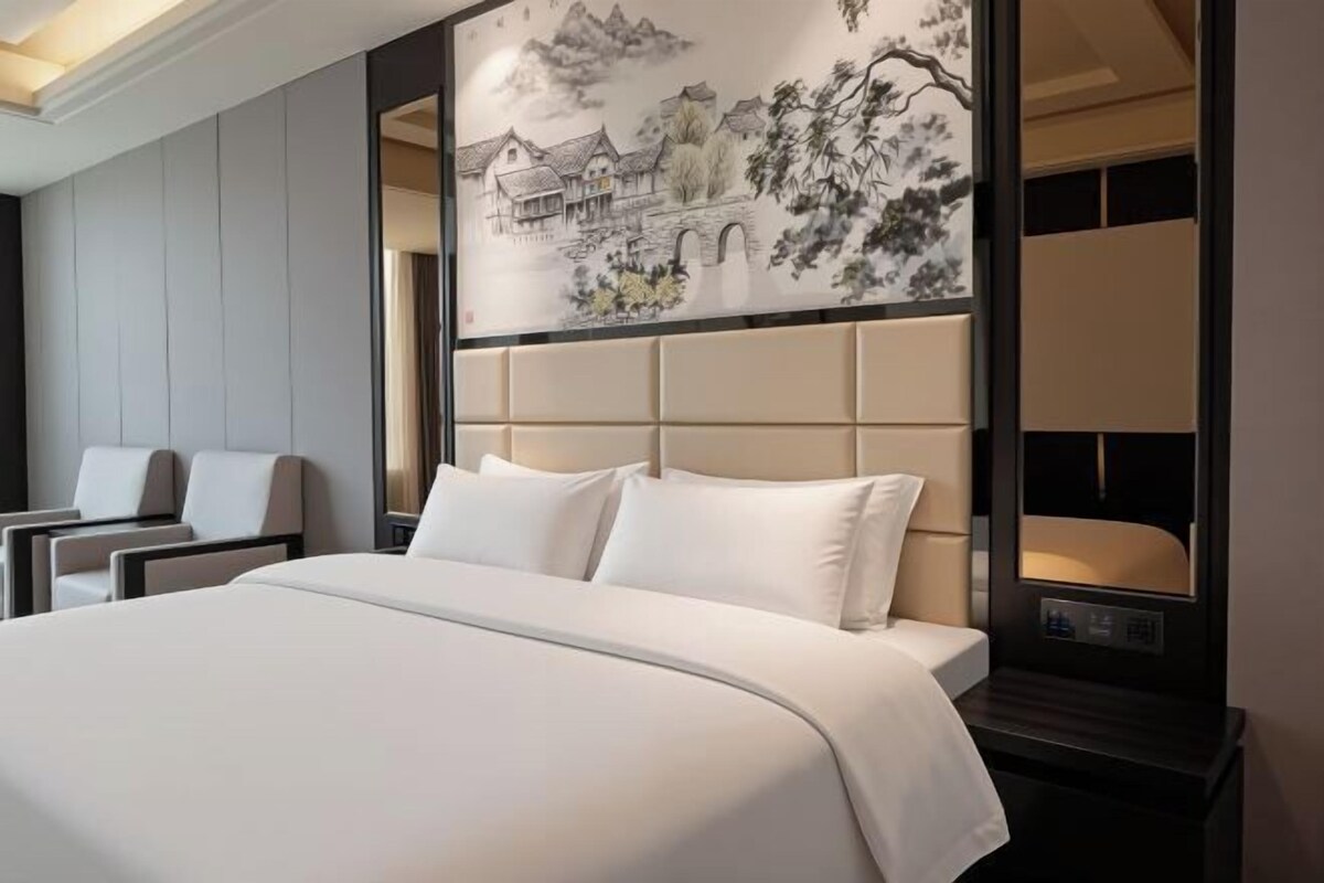 A hotel room with a large bed, a headboard with a mural, a desk, and two chairs.
