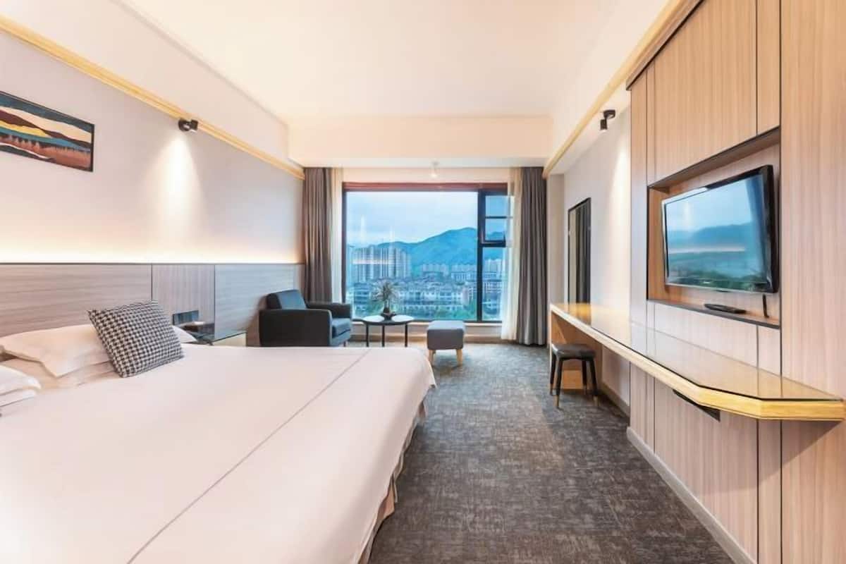 Quarto de hotel com uma cama grande, uma escrivaninha, uma televisão e vista para a cidade através de uma janela ampla.