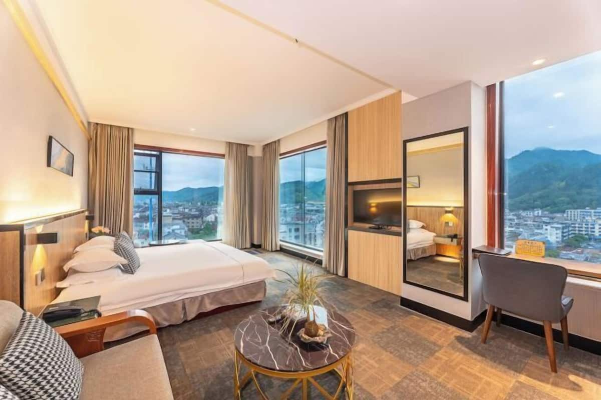 Quarto de hotel com cama, escrivaninha, cadeira, mesa redonda e uma grande janela com vista para a cidade.