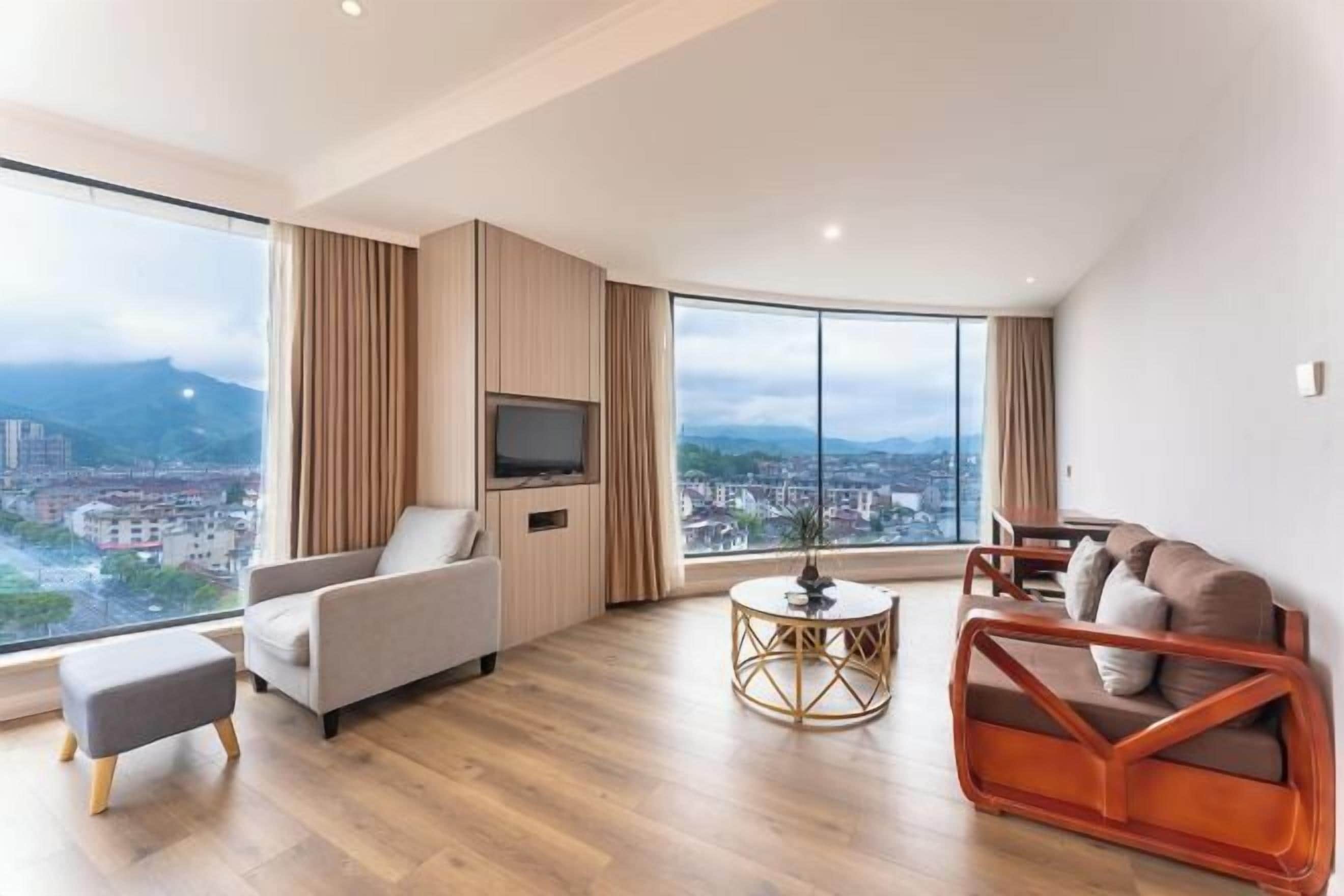 Un salon moderne avec un canapé, un pouf et une table basse ronde, de grandes fenêtres offrant une vue sur la ville, et un téléviseur à écran plat fixé sur un panneau en bois.