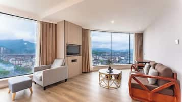 Un salon moderne avec un canapé, un pouf et une table basse ronde, de grandes fenêtres offrant une vue sur la ville, et un téléviseur à écran plat fixé sur un panneau en bois.