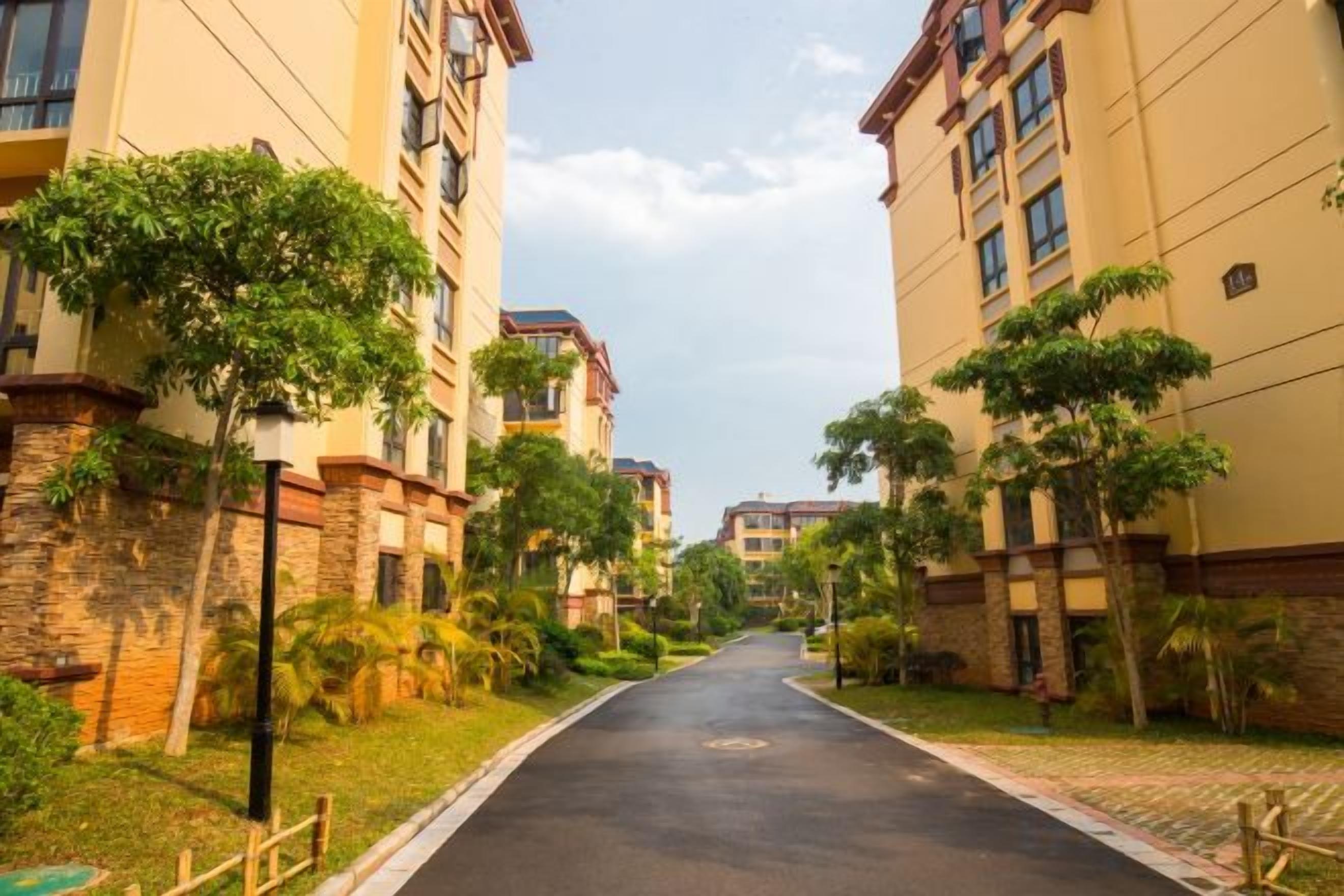 A residential street with multi-story buildings, a paved road, and greenery.