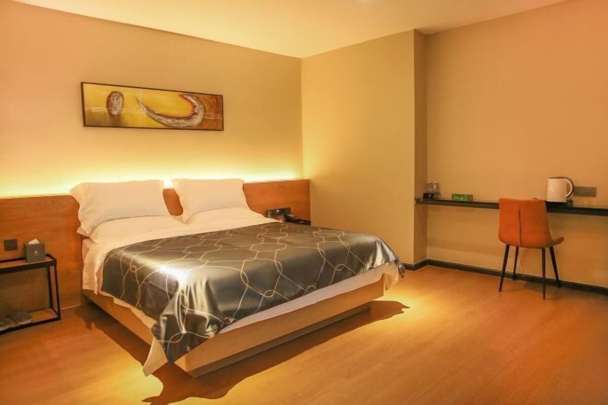 A modern hotel room with a bed, bedside table, chair, and a painting on the wall.