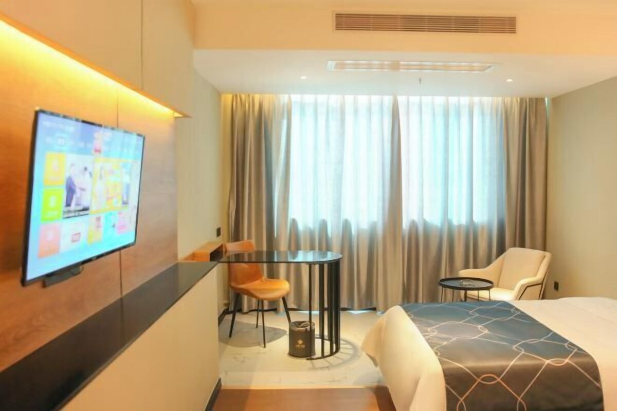 A hotel room with a bed, a TV mounted on the wall, a small table with chairs, and a window with curtains.