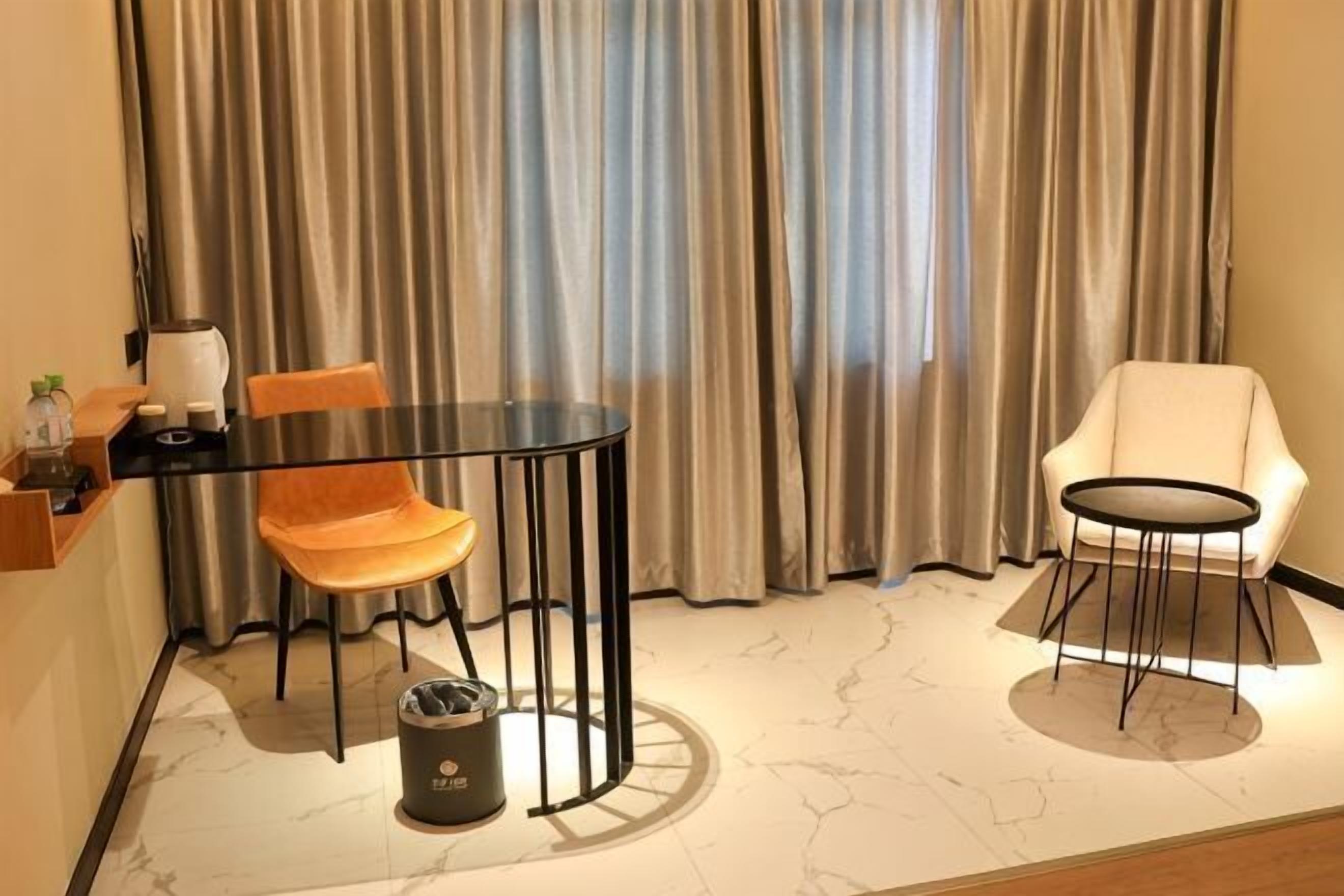 A room with a round table, two chairs, a trash bin, and a window with curtains.