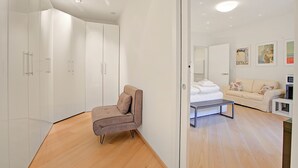 Une chambre d’hôtel moderne avec un plancher en bois, un canapé, un lit avec du linge de lit blanc, une petite table et une armoire blanche.