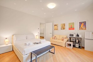 Et hotelværelse med seng, sofa, lille bord, bænk, tekøkken og loftlampe.