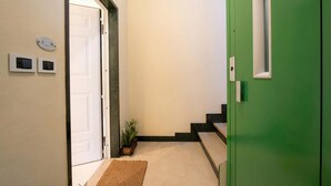 Un couloir avec une porte verte, une porte blanche, une petite plante en pot, un tapis à motifs et un interrupteur fixé au mur.