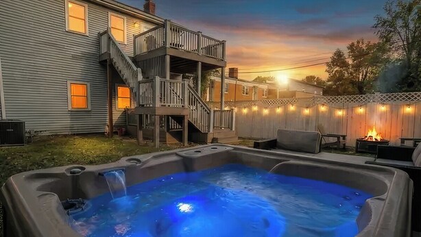 Un jacuzzi con cubierta de madera y escaleras, rodeado por un patio trasero con cerco y luces colgantes.
