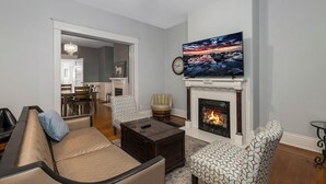 Una sala de estar con chimenea, un televisor montado encima y un comedor al fondo.