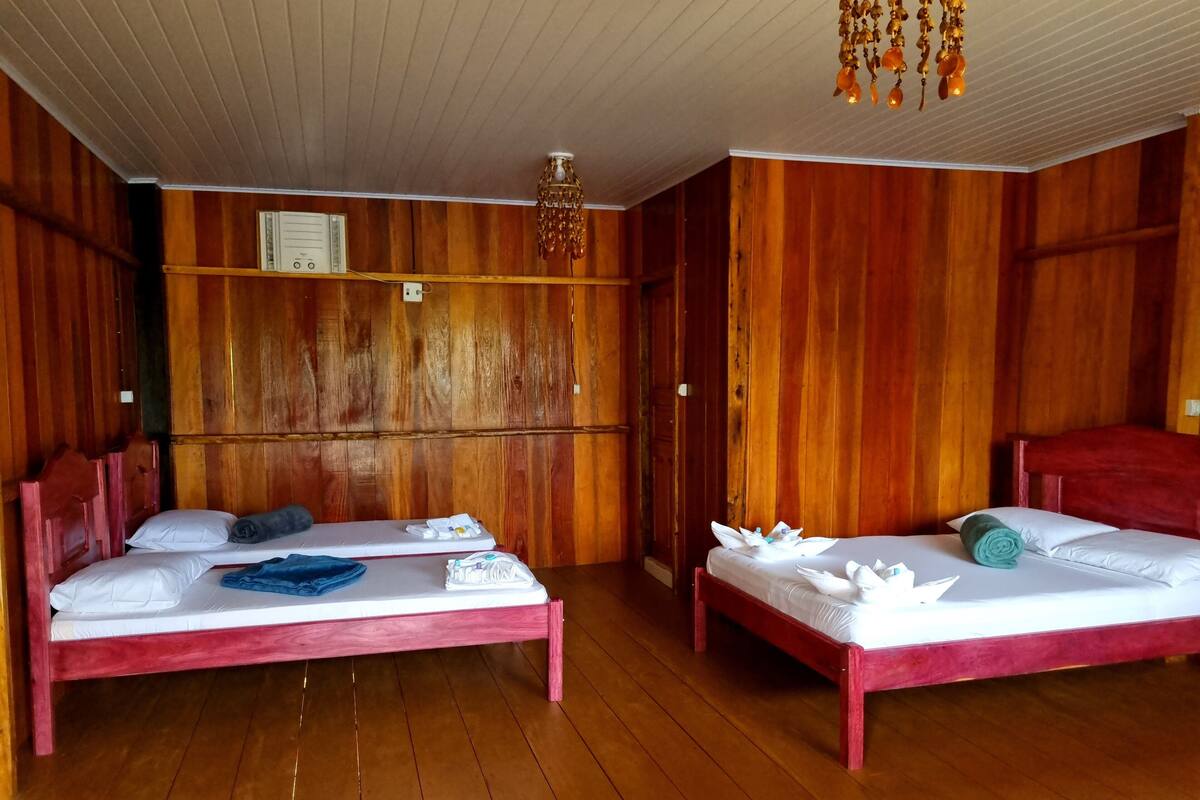 Ein Zimmer mit zwei hölzernen Betten, jeweils mit einem roten Kopfteil, weißen Laken und Handtüchern. Eine Holzzieldecke mit einer Pendelleuchte ist sichtbar.