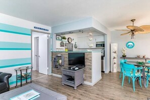 A modern living room with a TV, a small table, and a sofa. There is a kitchen area with a microwave and a dining table with chairs. - Mid Mari - Gorgeous Nautical Condo Near Beach, Shops & Restaurants- Condo 105 (Pompano Beach)