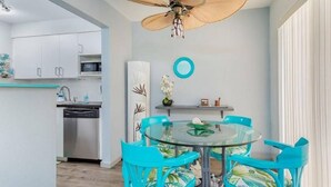 Une cuisine moderne avec des armoires blanches, un four à micro-ondes et un lave-vaisselle. On y trouve un coin repas avec une table en verre et des chaises turquoise. Un ventilateur de plafond avec un grand luminaire est visible au-dessus de la table.