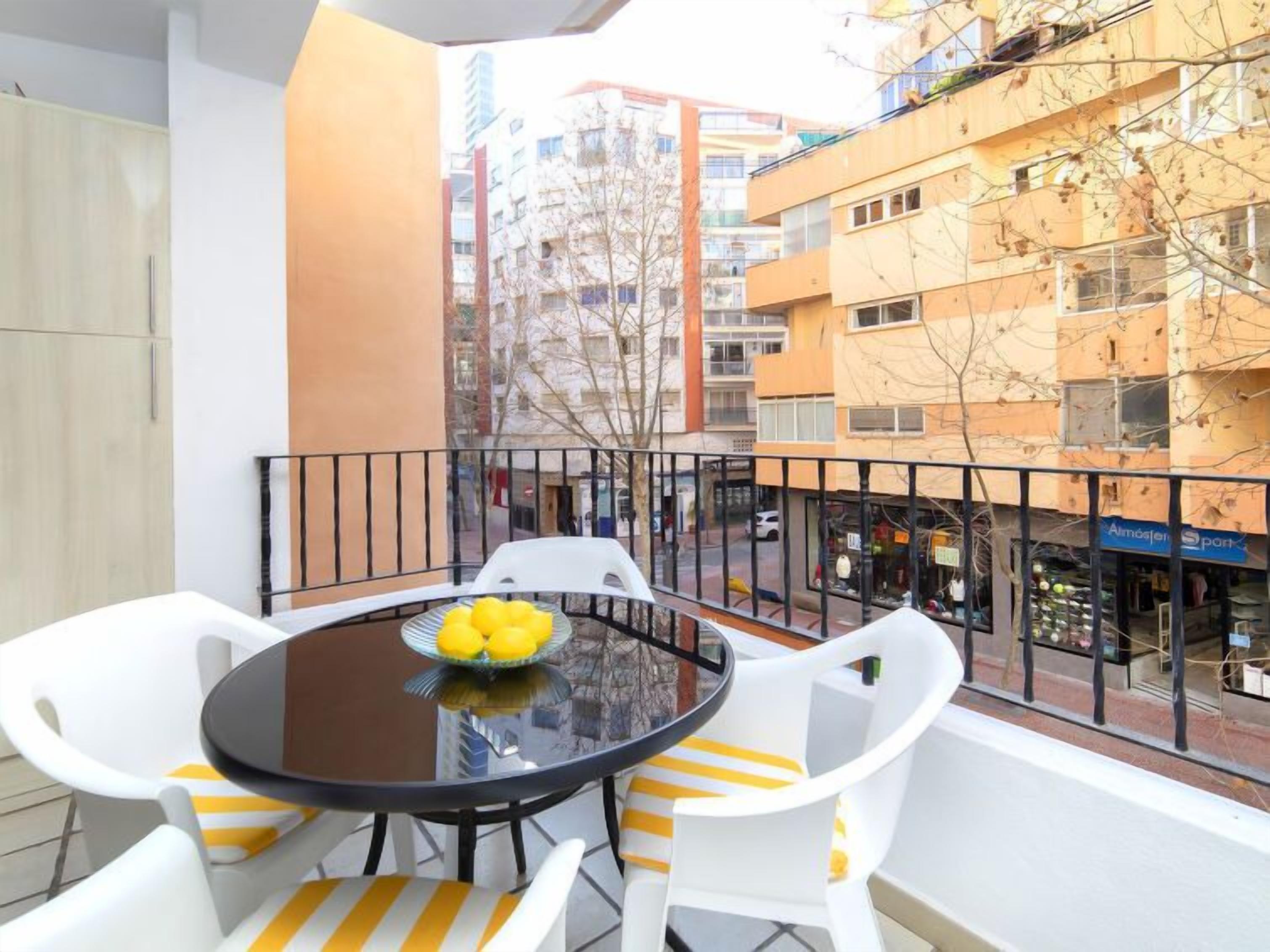 A balcony with a round table and white chairs, a bowl of lemons, and a view of a cityscape with buildings and shops.