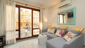 A living room with a grey sofa set, a small white side table, a bookshelf, and a large window with a view of a building and a balcony. - Eurosol by Interhome (Calpe/Calp)