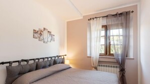 Un dormitorio con cama, ventana con cortinas translúcidas, radiador y cuadros en la pared.