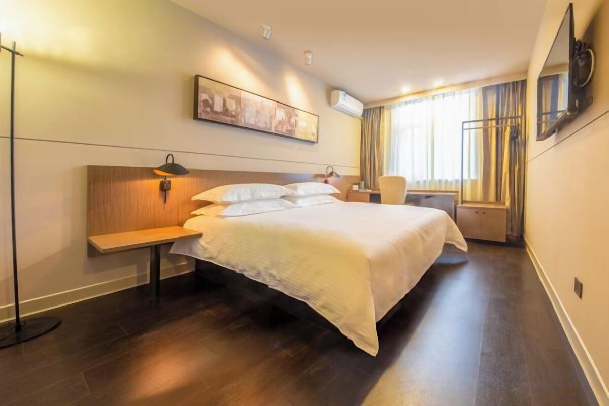 A hotel room with a large bed, bedside table, lamp, and a painting on the wall.