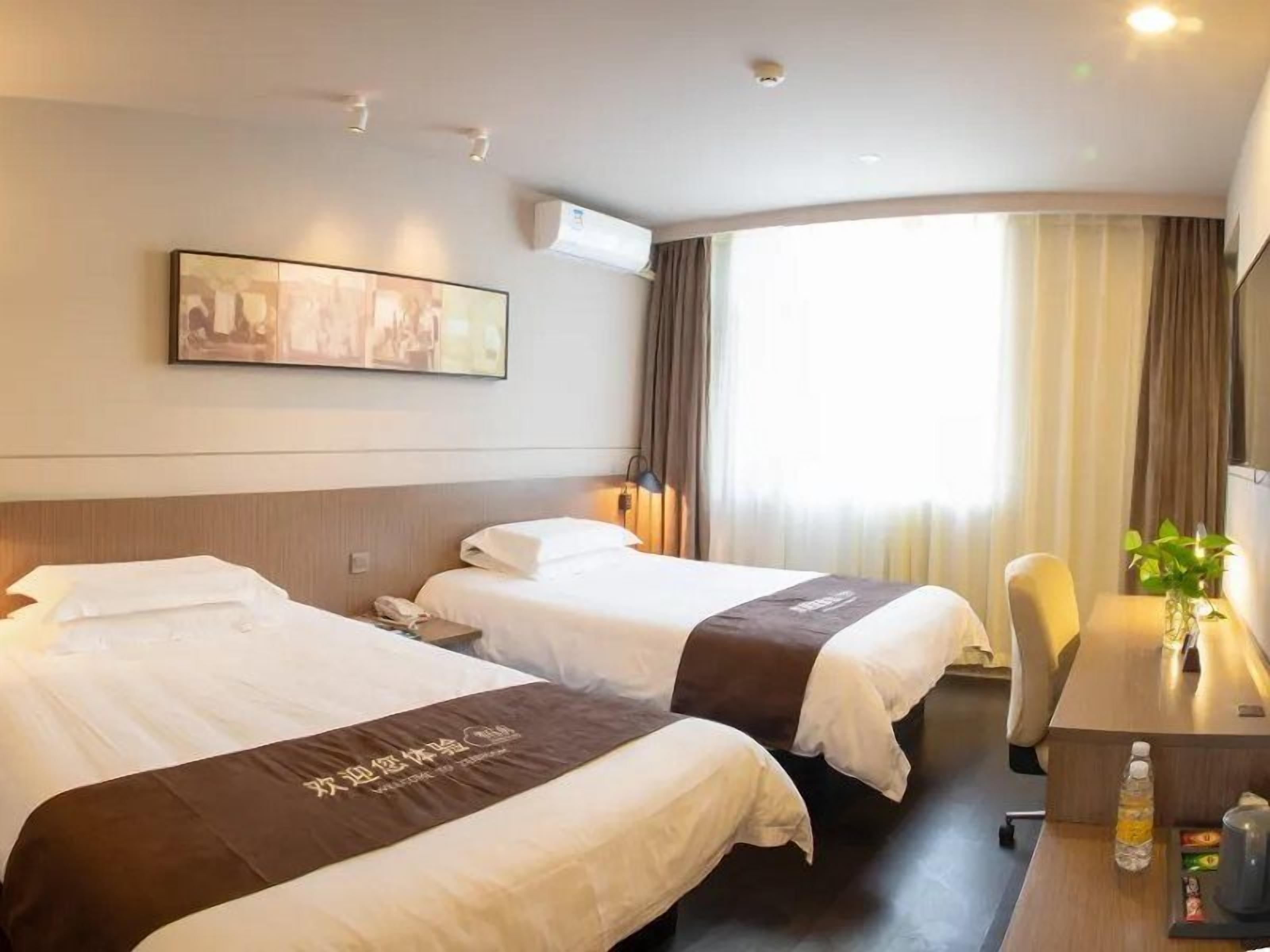 A hotel room with two beds, a desk, a chair, a window with curtains, and a wall-mounted air conditioner.