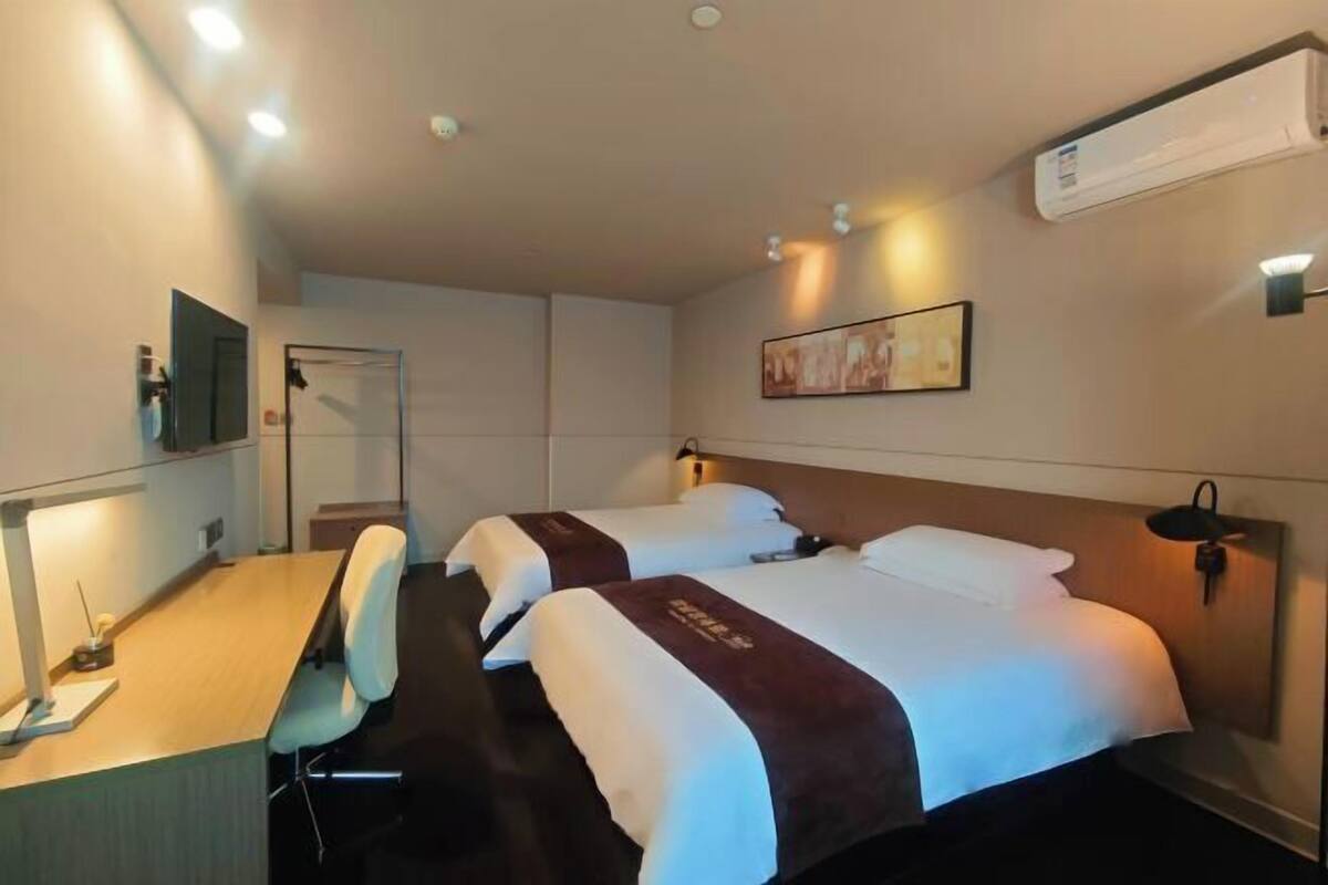 A hotel room with two beds, a desk, a chair, a mirror, and a wall-mounted air conditioner.