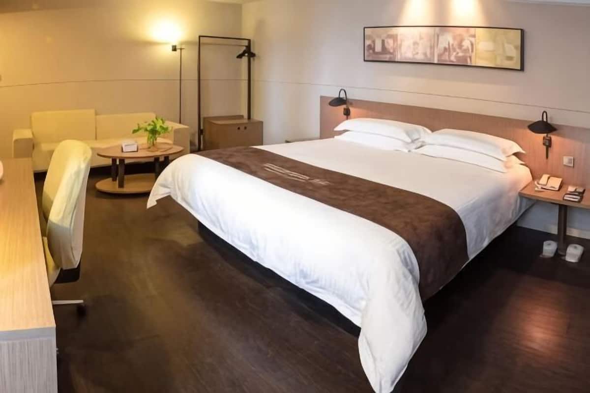 A hotel room with a bed, a desk, a chair, a small table with a plant, and a lamp.